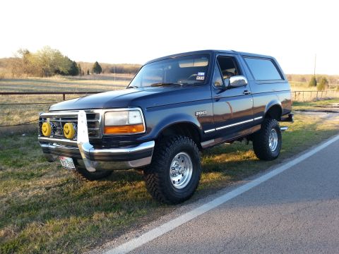 1995 Ford Bronco - This lifted blue 1995 Ford Bronco is parked on the side of a road with a rural landscape in the background.