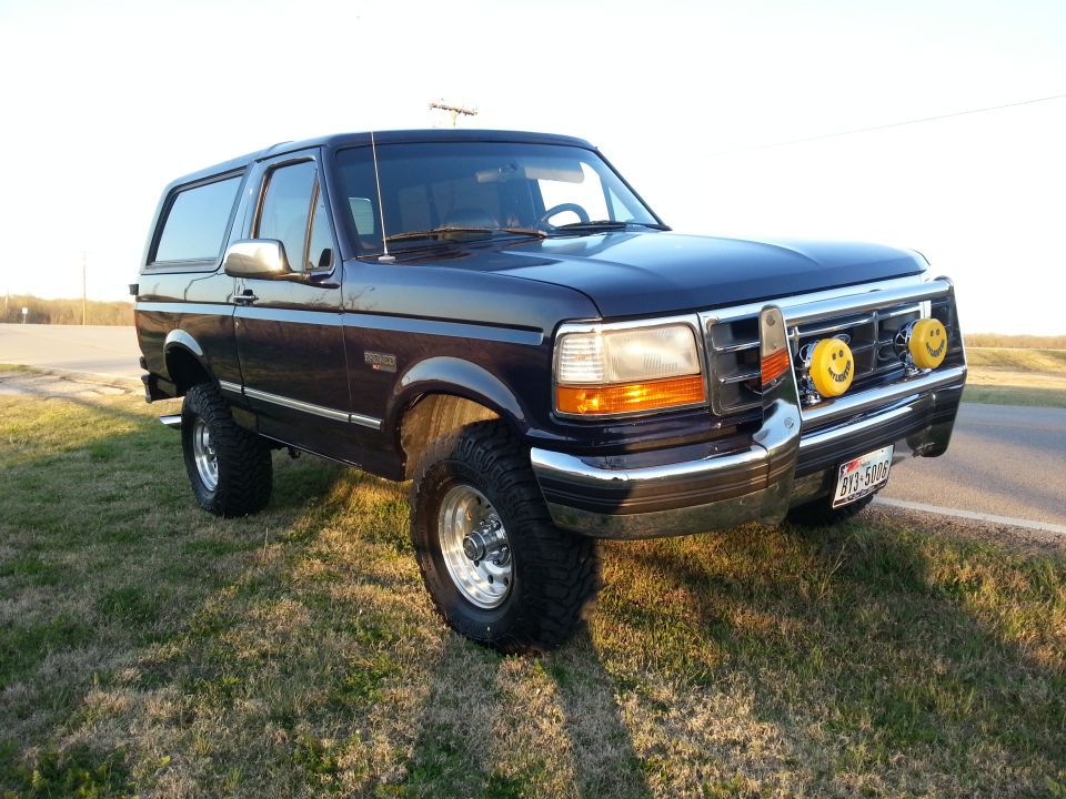 Dark blue 1995 Ford Bronco with smiley face fog lights and aftermarket wheels parked on grass