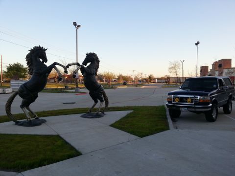 1995 Ford Bronco - A dark blue Ford Bronco is parked near two rearing horse statues under a clear sky.