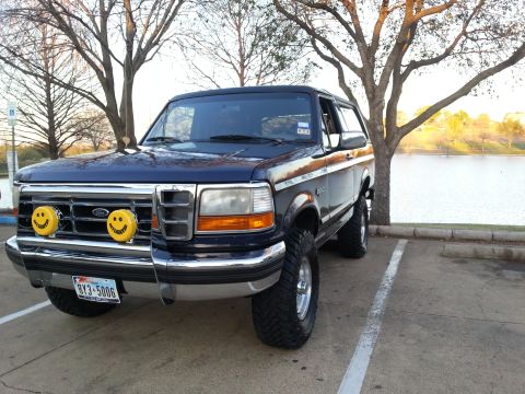 1995 Ford Bronco - A lifted 1995 Ford Bronco is parked in a lot near a body of water, with trees in the background.