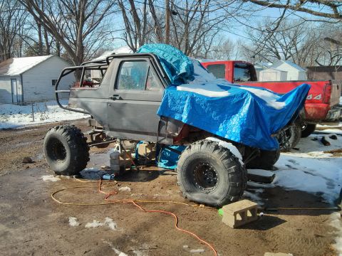 1986 Ford Bronco - This heavily modified SUV, possibly a Ford Bronco, sits outdoors with large off-road tires and a custom roll cage, partially covered by a b…