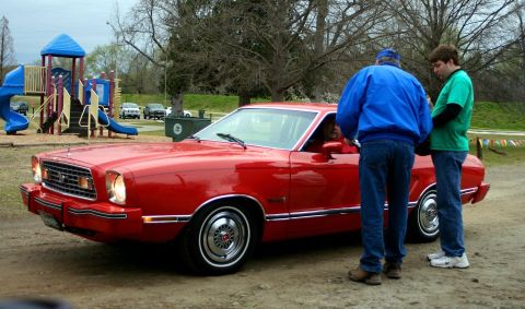 1980 Ford Mustang - A red 1975 Ford Mustang II coupe is parked near a playground while two men converse beside it.
