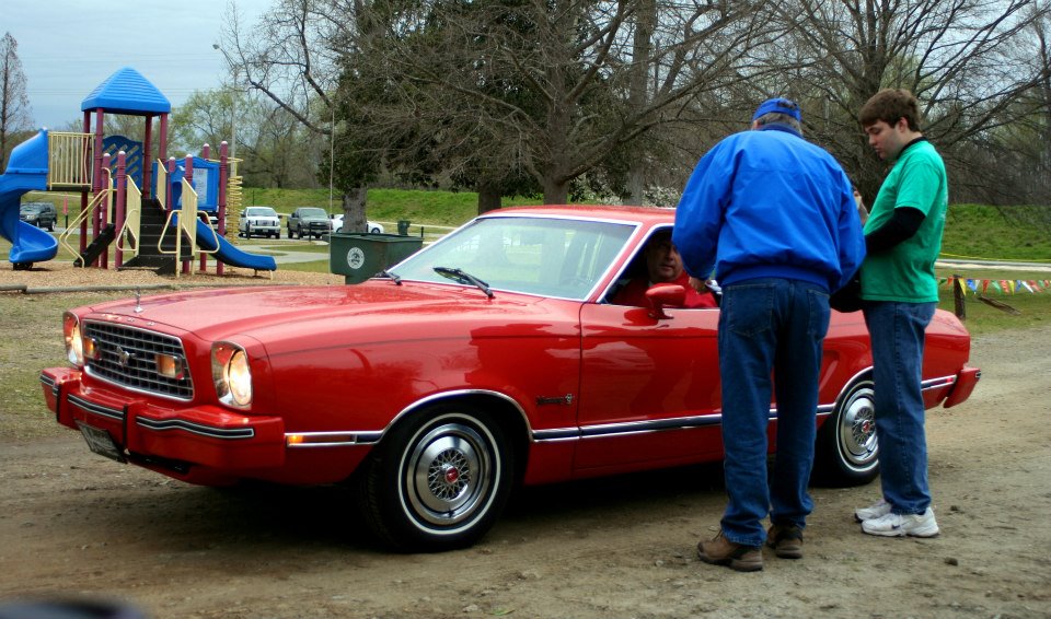 Red 1975 Ford Mustang II coupe parked at a playground with two men standing by the driver's door