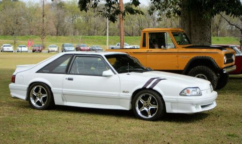 1980 Ford Mustang - A white Ford Mustang is parked on grass next to a yellow Ford Bronco, with other cars in the background.