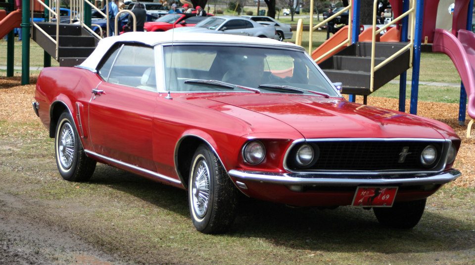 Red convertible classic car with white top parked on grass near playground equipment. License plate frame reads '70s 69'