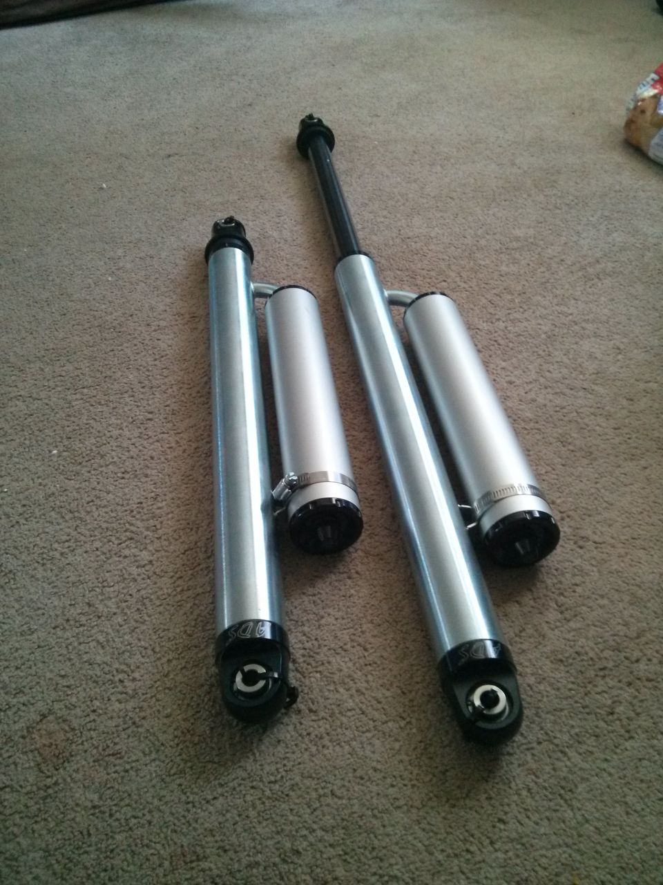 Two silver shock absorbers with black caps and clamps on a carpeted floor