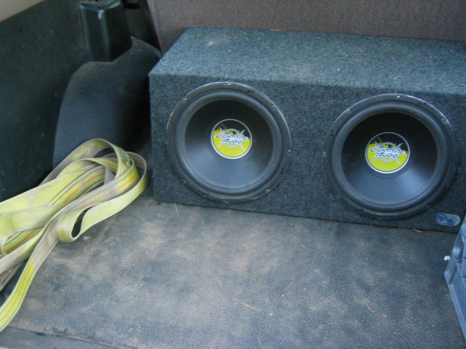 Gray carpeted dual Lightning Audio subwoofer box in a vehicle cargo area with a yellow strap beside it