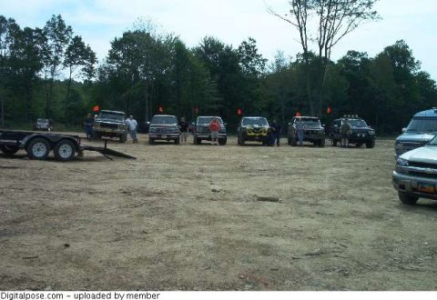 1993 Ford Bronco - Wide shot of a trail-run staging area: a row of six lifted 4x4 trucks and Broncos parked nose-out at the treeline, each with an orange whip…