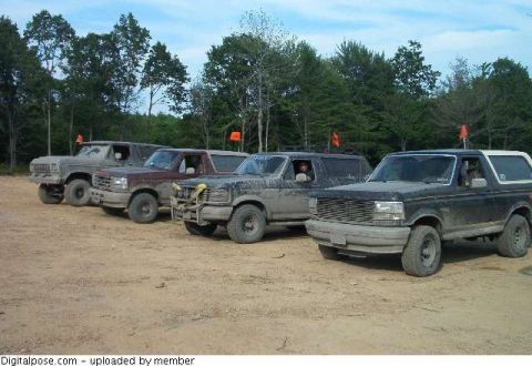 1993 Ford Bronco - Four mud-splattered full-size Ford Broncos and pickups lined up nose-out on a sandy trail staging area, each flying an orange whip flag. Th…