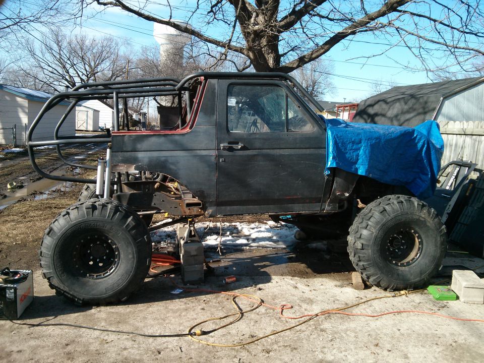 Custom off-road SUV with large tires, roll cage, and tarped front end, sitting on blocks