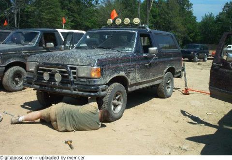 1993 Ford Bronco - A mud-covered dark blue 1987-91 Ford Bronco propped up with an orange hi-lift/bottle jack under the rear while a person lies on the sand wo…