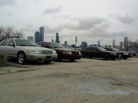 2003 Ford Crown Victoria - Several Ford Crown Victorias are parked in a lot with a city skyline in the background.