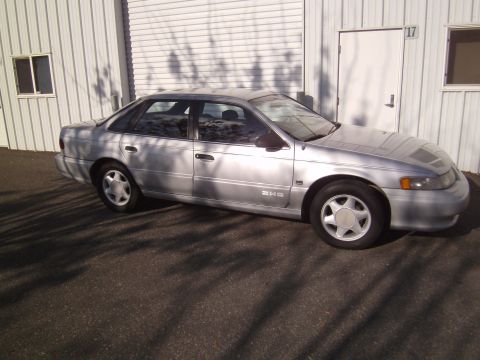 1992 Ford Taurus - A silver 1992 Ford Taurus sedan is parked on asphalt in front of a corrugated metal building.