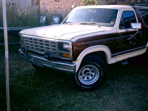 1980 Ford Bronco - Front three-quarter view of a two-tone brown and cream 1980s Ford Bronco parked on grass next to a brick building, with an eggcrate grille,…