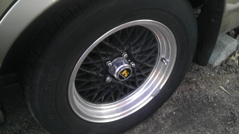 Close-up of a car wheel with a black mesh design and a silver lip. Center cap has a yellow and black logo