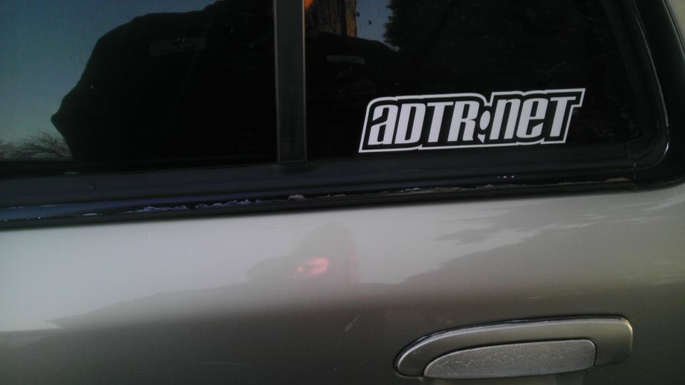 Silver car door with a chrome handle and a black window sticker reading ADTR.net. Watermark "SUPERMOTORS.NET" visible