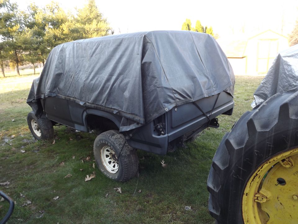 Tarp-covered two-door SUV body shell on lifted suspension parked on grass beside a tractor tire