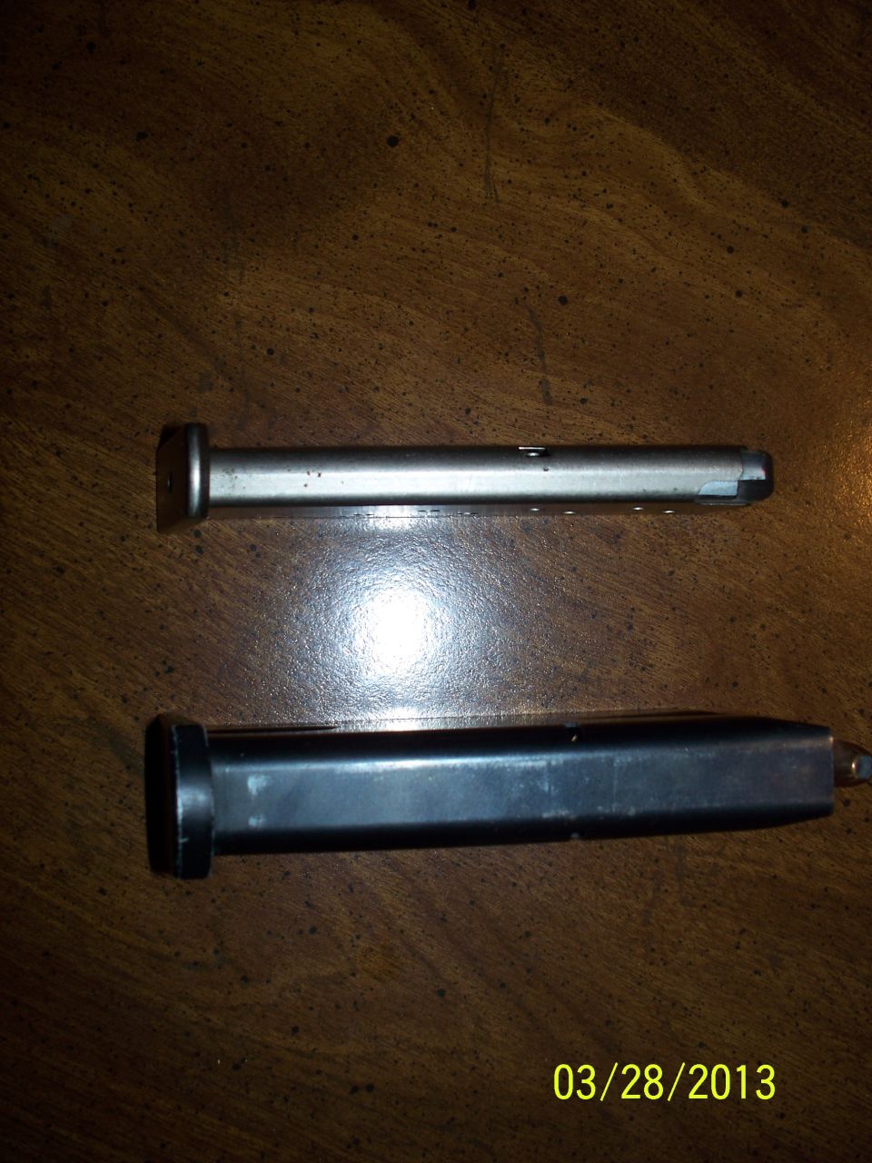 Slim stainless pistol magazine above a wider blued double-stack magazine on dark wood
