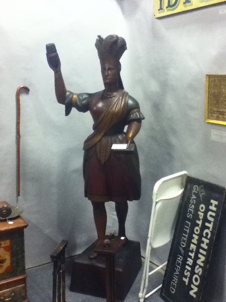 Wooden statue of a woman holding a glass and paper, with a cane and sign in background