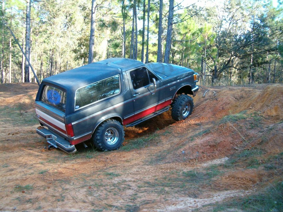 Dark gray Ford Bronco with red stripe, lifted on off-road tires, climbing a dirt hill