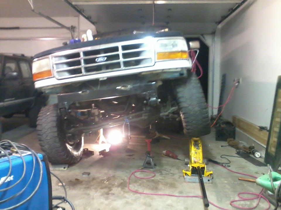 1992 Ford Bronco front end lifted on jack stands in a garage. Another SUV is visible to the left
