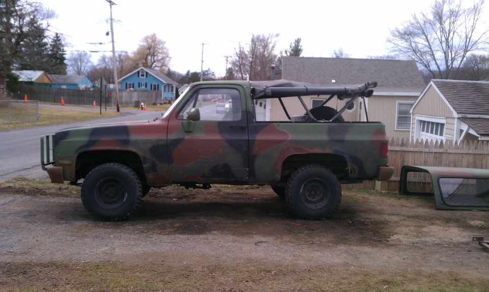 Camouflage pickup truck with a soft top removed, parked on a dirt lot next to a wooden fence