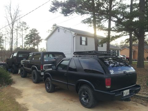 1986 Ford Bronco - Multiple black SUVs are parked in a line on a dirt driveway next to a house.