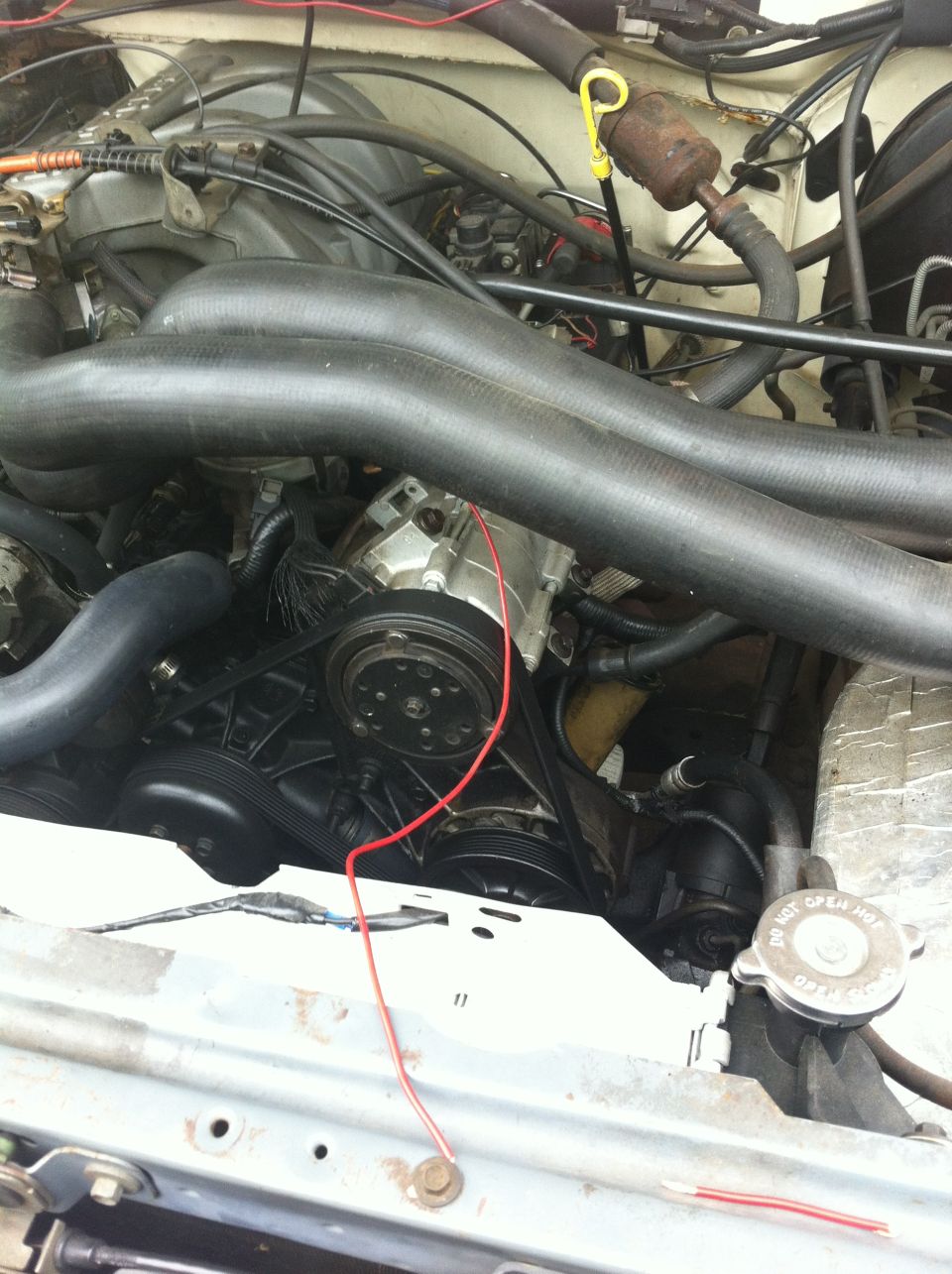 Engine bay with hoses, belts, and engine components visible. A red wire runs across the engine