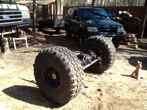 1989 Ford Bronco - Axle assembly with large tires sits in front of a dark green SUV and a trailer.