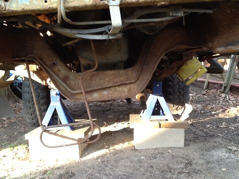 1989 Ford Bronco - The underside of a vehicle is supported by jack stands, revealing rusted frame components and suspension parts.