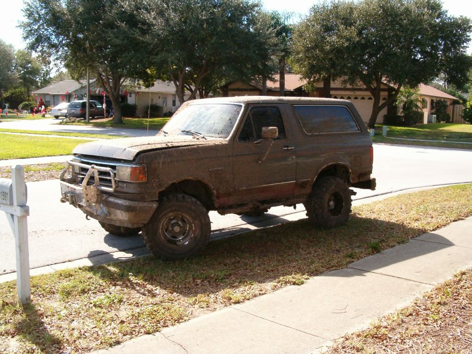 Mud-covered brown Ford Bronco with aftermarket bumper parked on grass, 3/4 front view