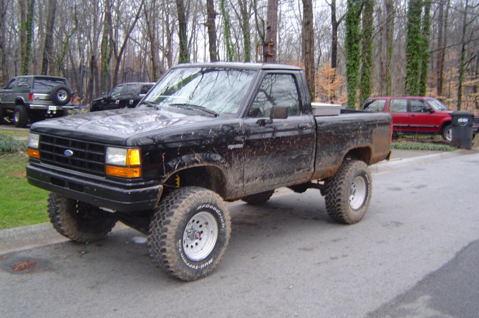 1989 Ford Ranger pickup truck, lifted with off-road tires, parked on a street with trees in the background. Another SUV and a red car are visible
