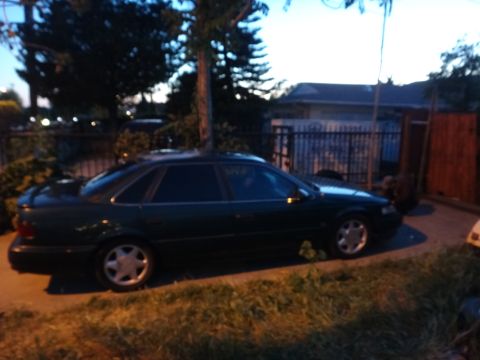 1993 Ford Taurus - A dark green sedan is parked outdoors at dusk, with a fence and trees in the background.