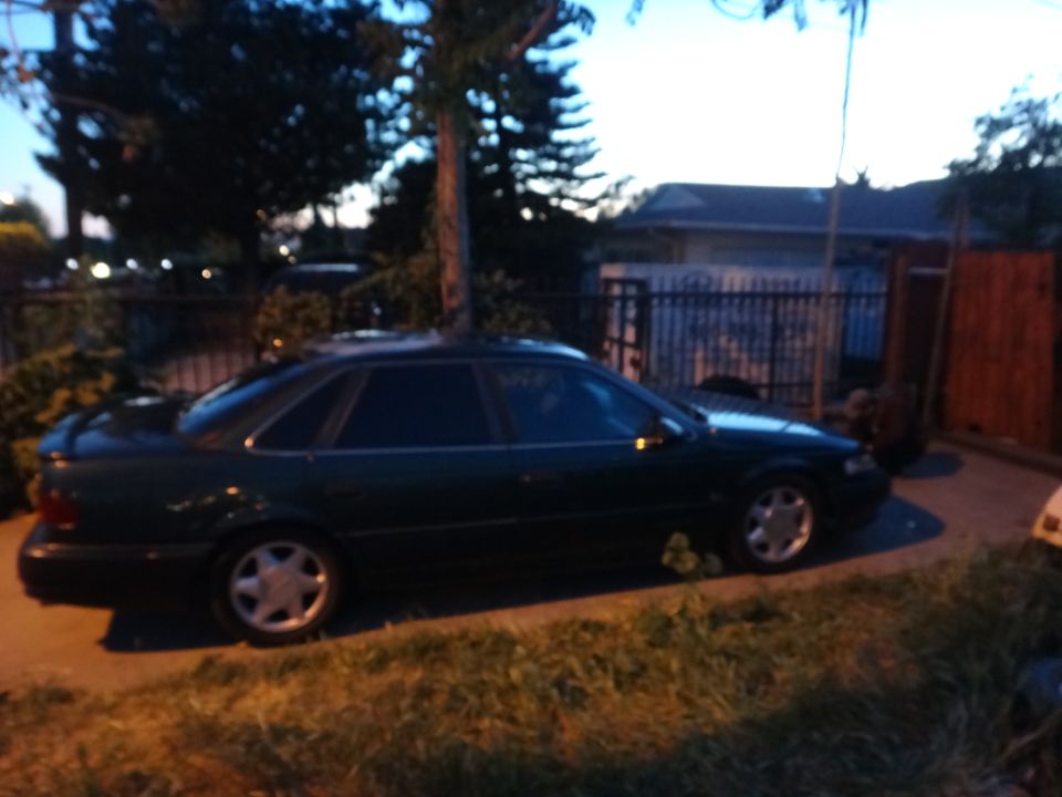 Dark green sedan parked on a grassy area, side view. The car has a spoiler and alloy wheels