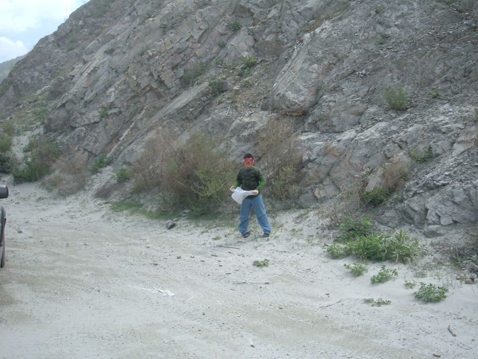 Person stands in a dirt area with a rocky hillside behind them. A vehicle is partially visible on the left