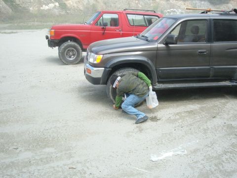 1988 Ford Bronco - A person crouches to inspect the tire of a dark SUV, with a red Ford Bronco parked behind it.