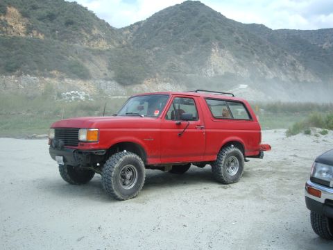 1988 Ford Bronco - This red 1988 Ford Bronco is seen from a 3/4 front angle, parked in a dusty, outdoor setting with mountains visible.