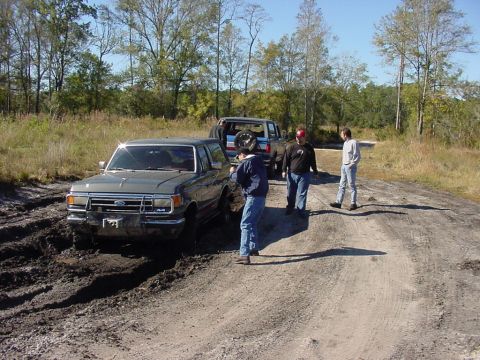 1991 Ford Bronco - A gray 1991 Ford Bronco is stuck in the mud while onlookers stand nearby on a dirt road.