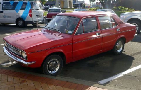 1969 MINI Cooper - A red sedan is parked in a lot next to other vehicles under a bright sun.