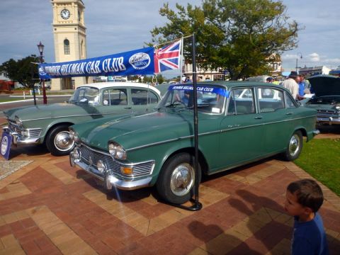 1969 MINI Cooper - A green Humber Hillman sedan is parked next to another vintage car, with a club banner and a clock tower in the background.