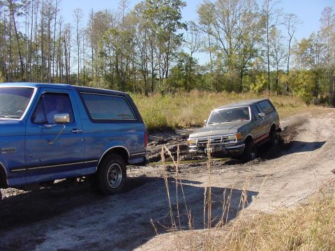 1991 Ford Bronco - Two Ford Broncos navigate a muddy off-road trail, with one vehicle stuck in the mire.