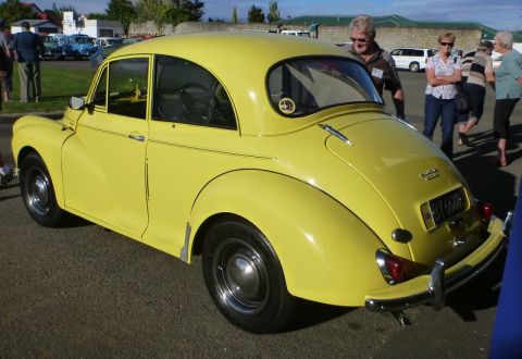 1969 MINI Cooper - A yellow Morris Minor is displayed at an outdoor event with several people observing.