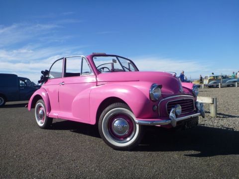 1969 MINI Cooper - A pink Morris Minor convertible is parked on asphalt under a blue sky with clouds.