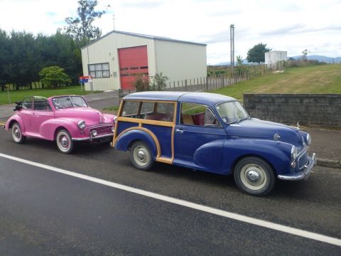 1969 MINI Cooper - Two classic Morris Minors, a pink convertible and a blue woody wagon, are parked on the side of a road.