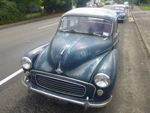 1969 MINI Cooper - A blue Morris Minor 1000 is parked on the side of a road, with other vehicles in the background.