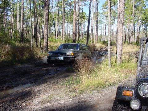 1991 Ford Bronco - A Ford Bronco navigates a muddy trail in a wooded area, with another off-road vehicle nearby.