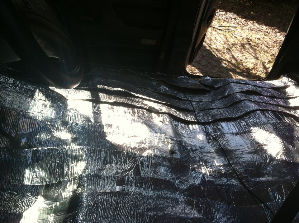 Interior of a vehicle with silver soundproofing material covering the floor and lower walls. Sunlight streams in from an open door
