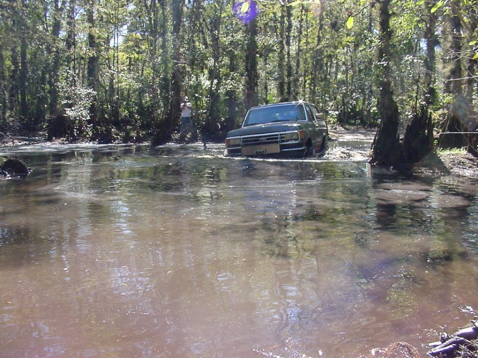 Dark green 1991 Ford Bronco driving through water in a wooded area