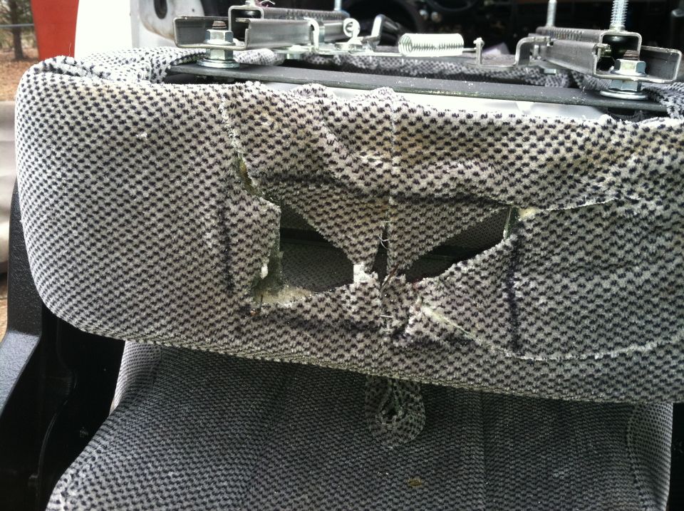 Torn fabric on a vehicle seat cushion, showing foam underneath. Metal brackets are visible above