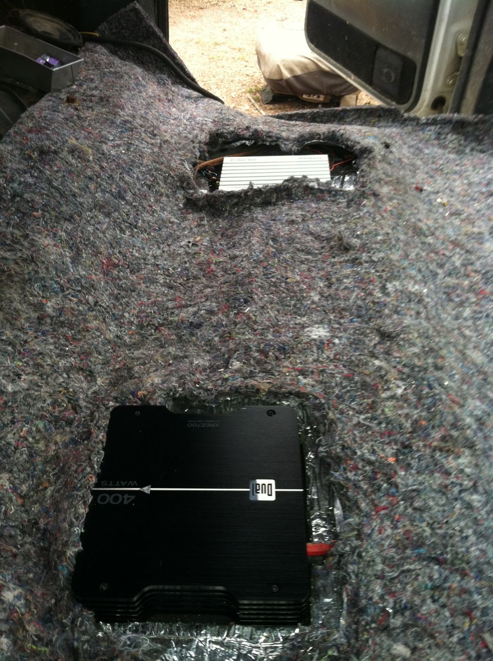 Interior view of vehicle with carpet removed, showing two audio amplifiers installed